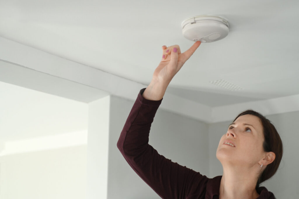 Young adult woman checks a smoke detector on a ceiling, representing the CPSC's smoke detectors warning