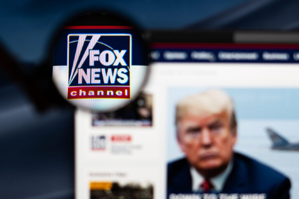 Fox News website homepage. Fox News channel logo visible through a magnifying glass, representing the Fox election fraud lawsuit.