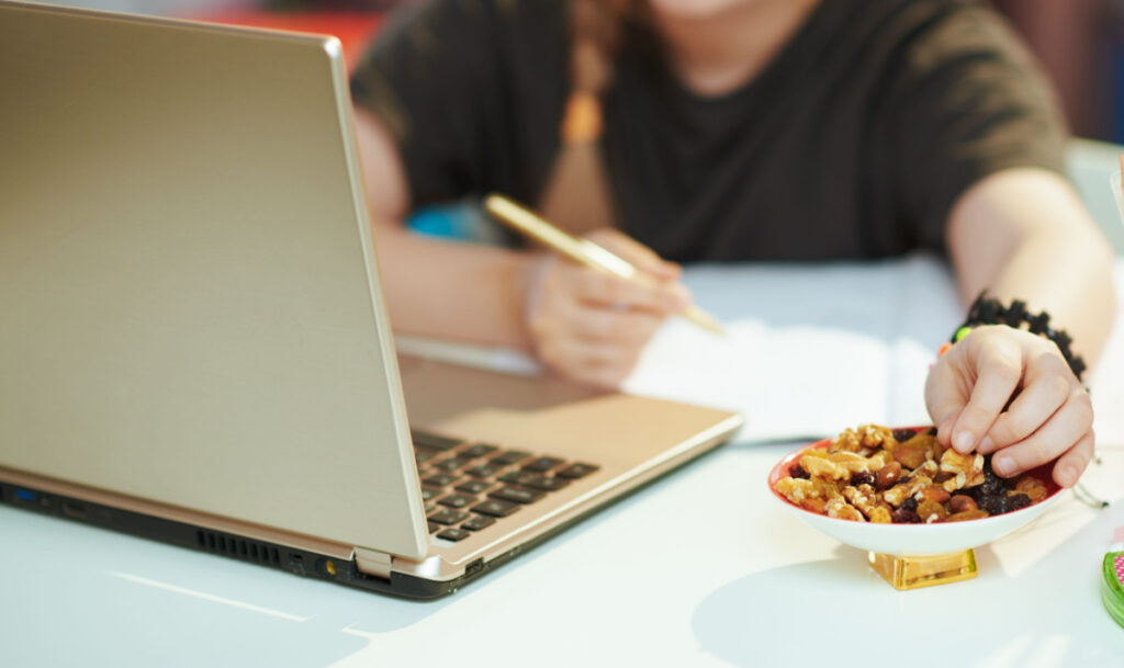 Closeup on girl in grey shirt with laptop homeschooling and eating nuts.