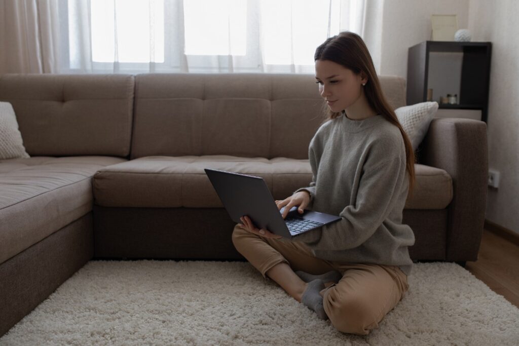 A woman sits cross-legged on the floor in front of a sofa, smiling while using a laptop, representing the class action settlements closing in July.