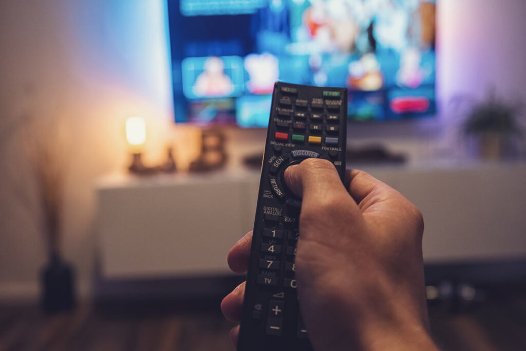 Close-up of a man's hands pointing a remote toward a TV, representing FCC cable and satellite pricing requirements.