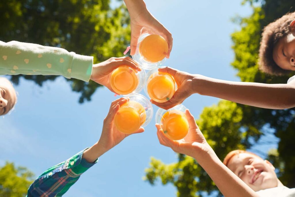 Low angle of kids holding glasses of orange juice against the sky, representing orange juice prices.