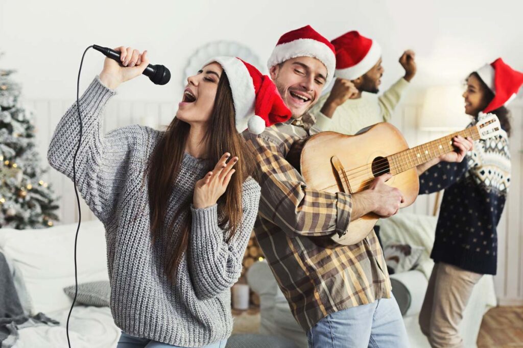 A young couple wearing Christmas hats singing and playing guitar, representing the wallet-friendly holiday activities.