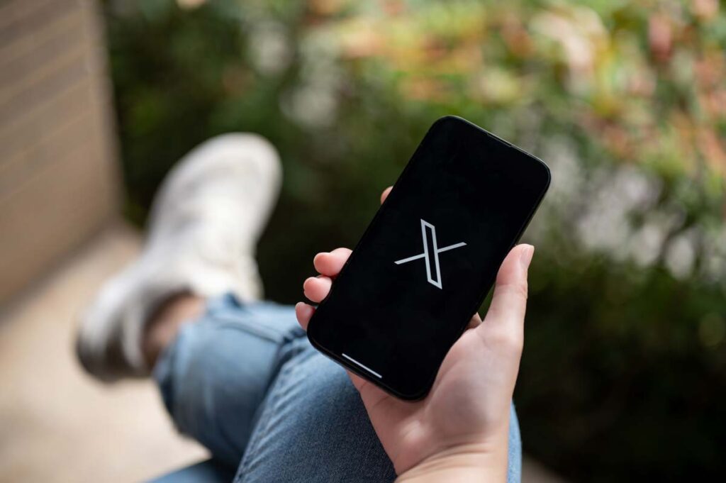 Close up of a woman holding a smartphone with the X logo displayed, representing the X advertiser loss Media Matters lawsuit.