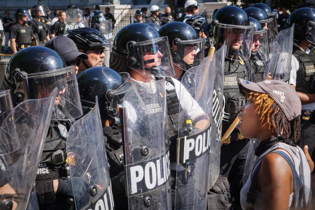 A protestor standing in front of a line of armed police officers at a protest, representing the New York George Floyd protests class action lawsuit settlement.