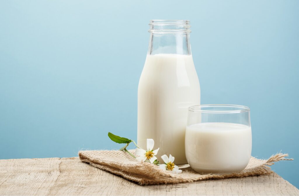 A glass bottle of milk sits next to a short glass of milk, sitting with small white flowers on wooden table against a blue background, representing the Lyons Magnus class action lawsuit settlement.