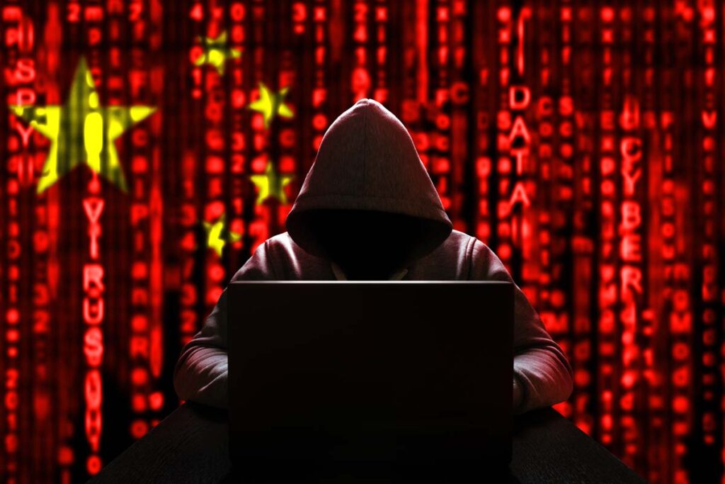 A hooded hacker using a computer with a Chinese flag in the background, representing Chinese hackers targeting United States systems.
