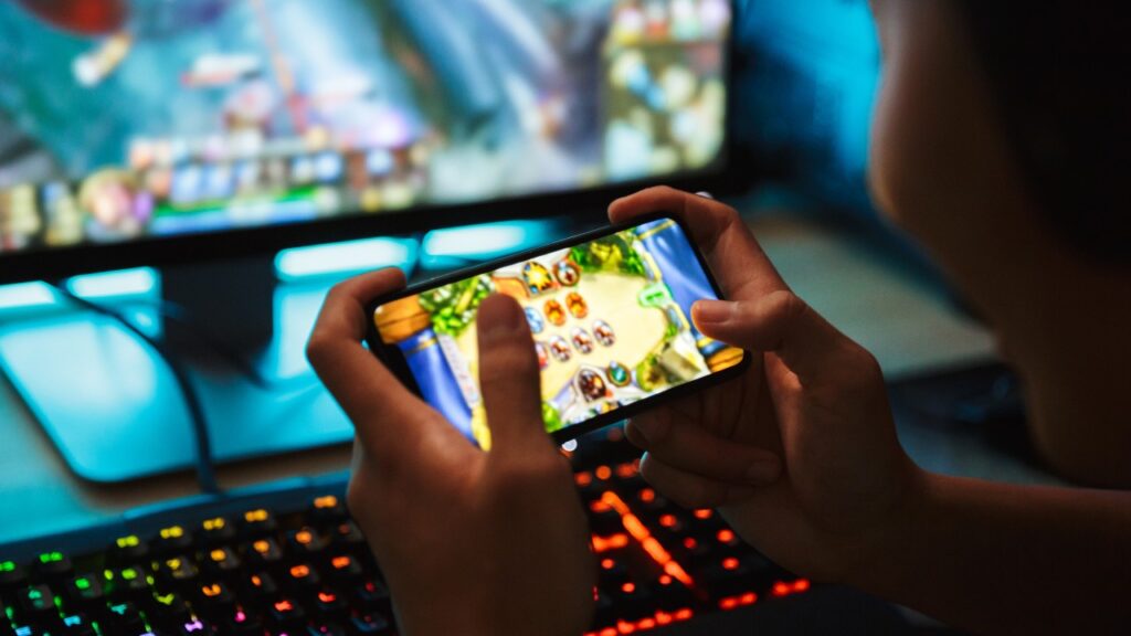 Teenage gamer boy wearing headphones and using a backlit colorful keyboard to play video games on his smartphone and computer in a dark room