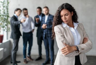 Workplace Sexism And Bullying. Unhappy Victimized Businesswoman Standing While Her Male Colleagues Whispering Behind Her Back