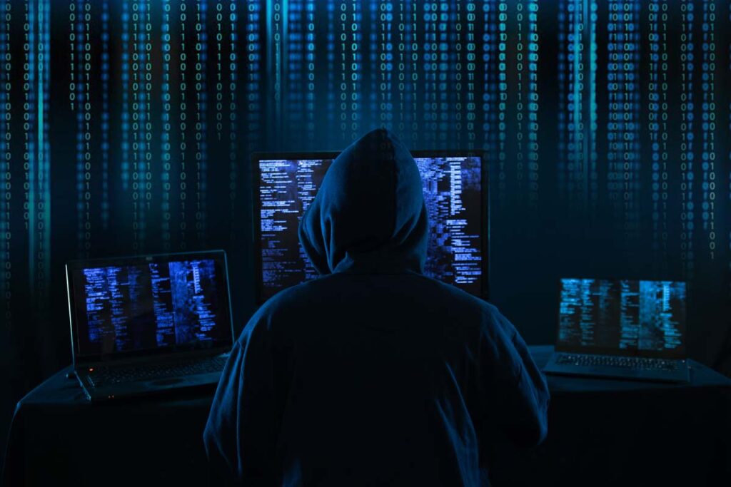 Back view of a hooded hacker using a computer, with a binary code overlay, representing the auto dealers data breach.