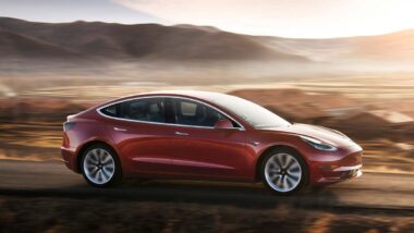 A red Tesla model 3 driving along a roadway, representing the Tesla recall.