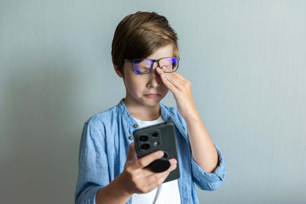 A young boy rubbing his eyes while using a smartphone, representing social media warning labels.