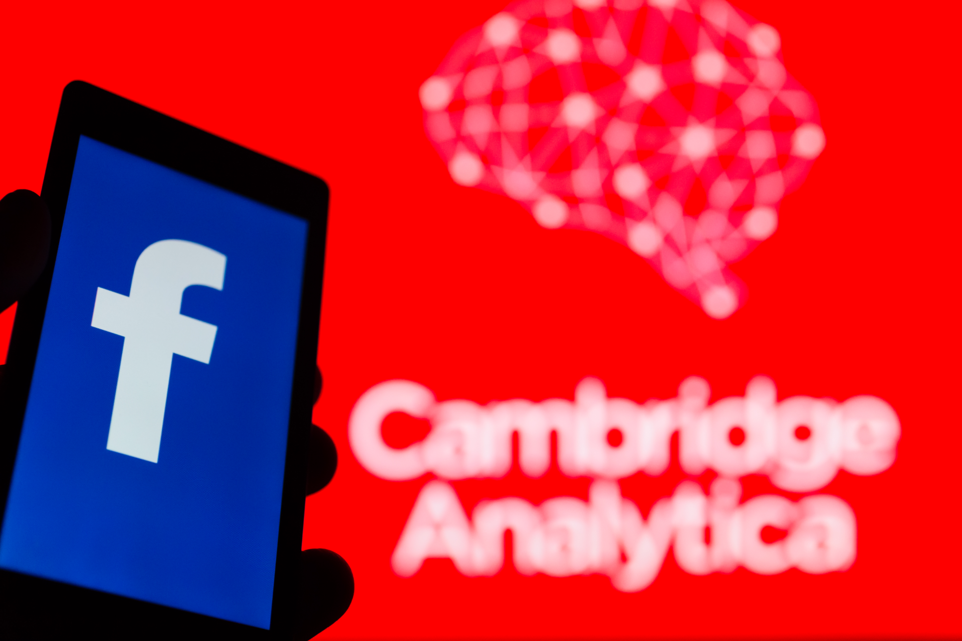 A hand holds a phone with showing a facebook logo. The Cambridge Analytica white-on-red logo is blurry in the background.