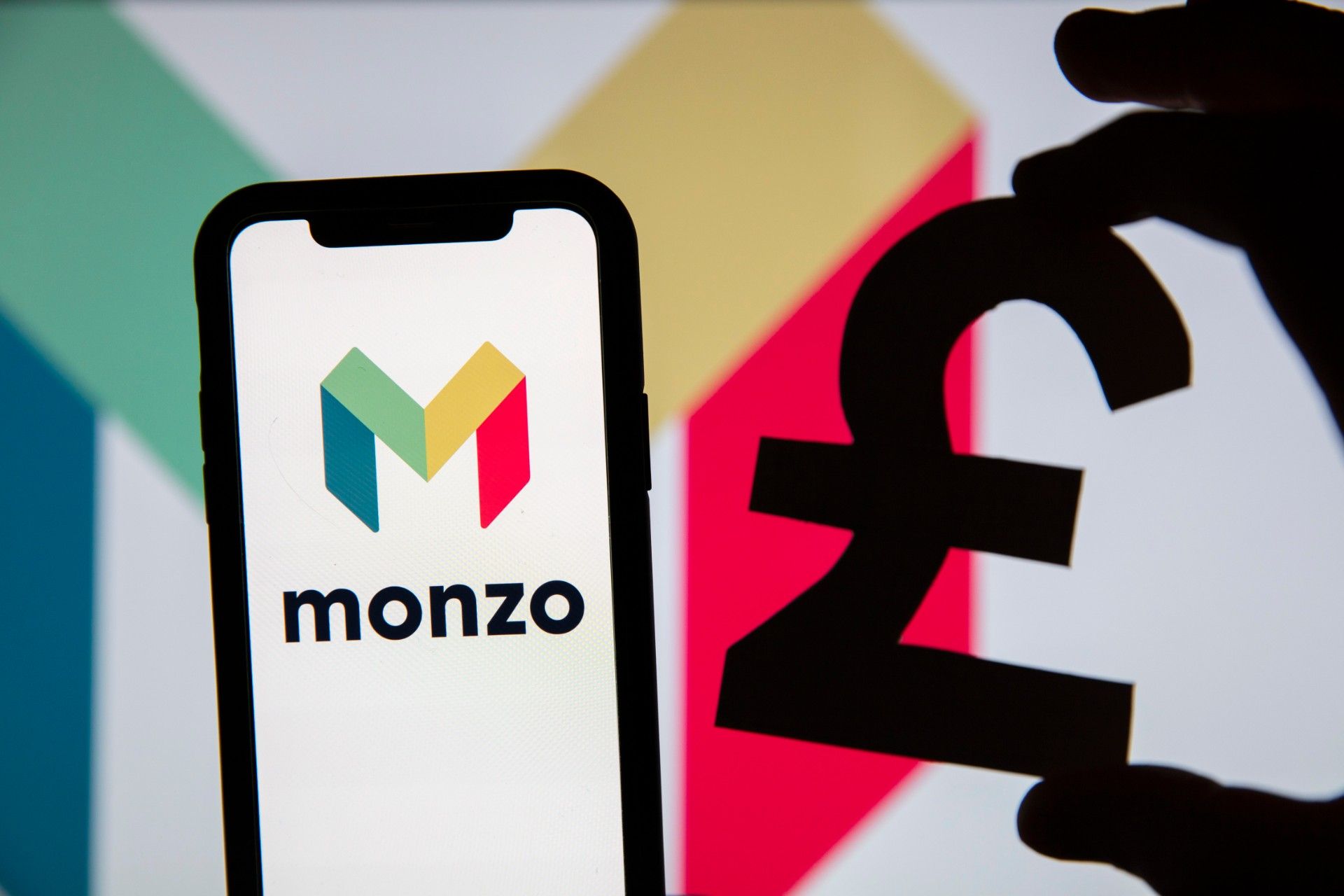 Monzo logo on smartphone and silhouette of a hand holding a pound symbol in front of Monzo background