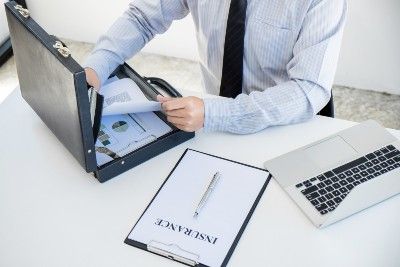 Man puts paperwork in briefcase on desk next to insurance paper on clipboard and laptop - insurance premiums