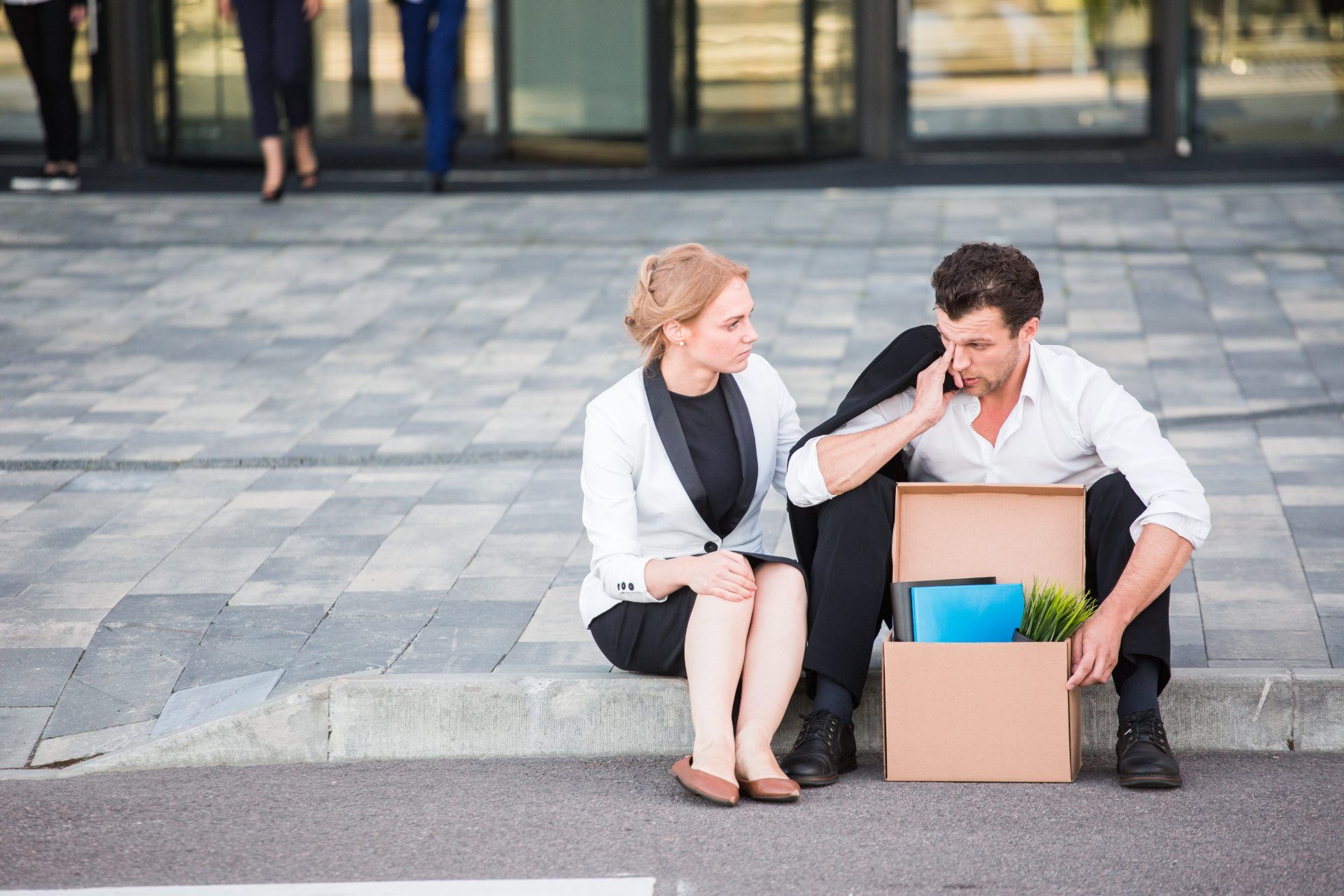 Woman comforts man sitting on curb with box of office items - redundancies