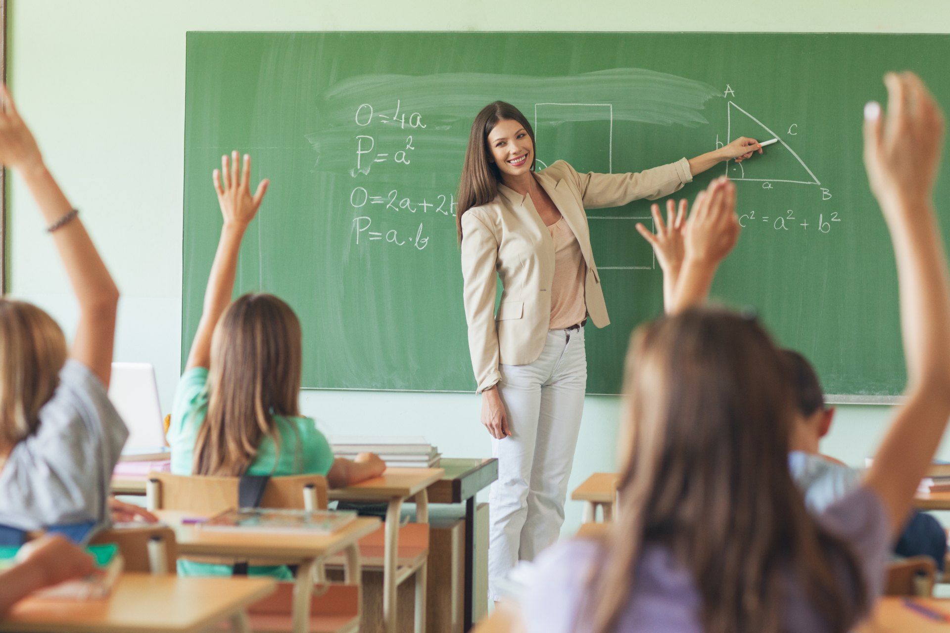A female teacher teaches math at chalkboard as students raise hands - pay rises