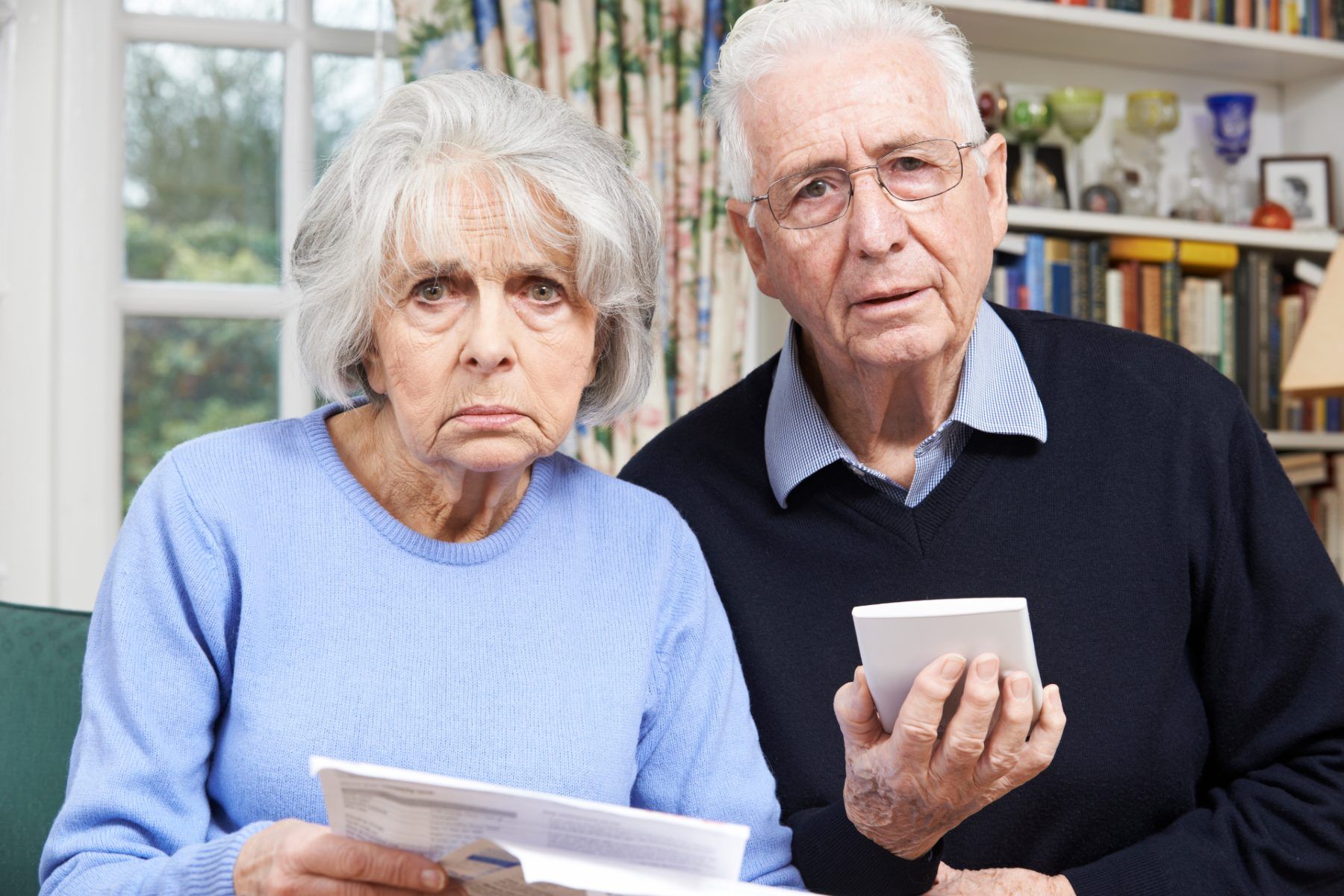 A worried older couple holds paperwork and a calculator while looking into the camera - pension scams