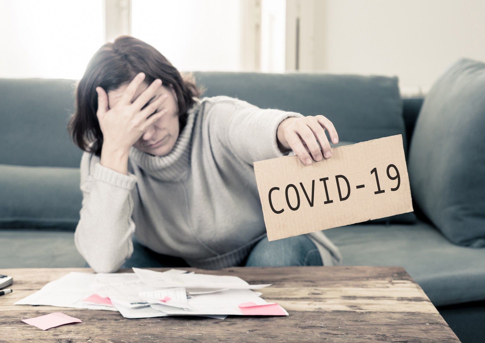 Woman stressed over finances sits on her sofa while holding out a sign that says "COVID-19" - payment deferrals