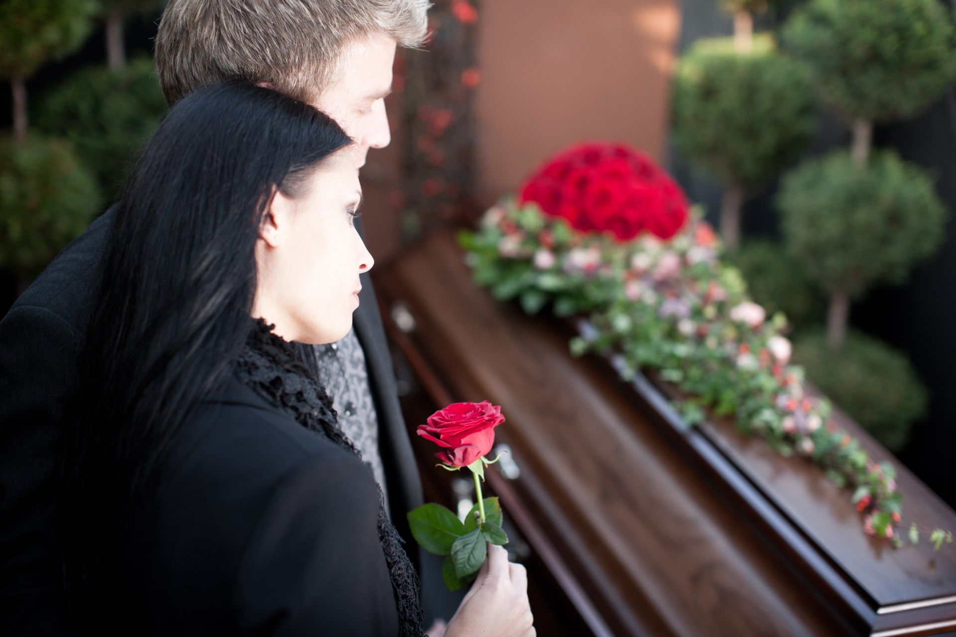 Mourners stand near a brown casket with red roses on top during a funeral - funeral costs