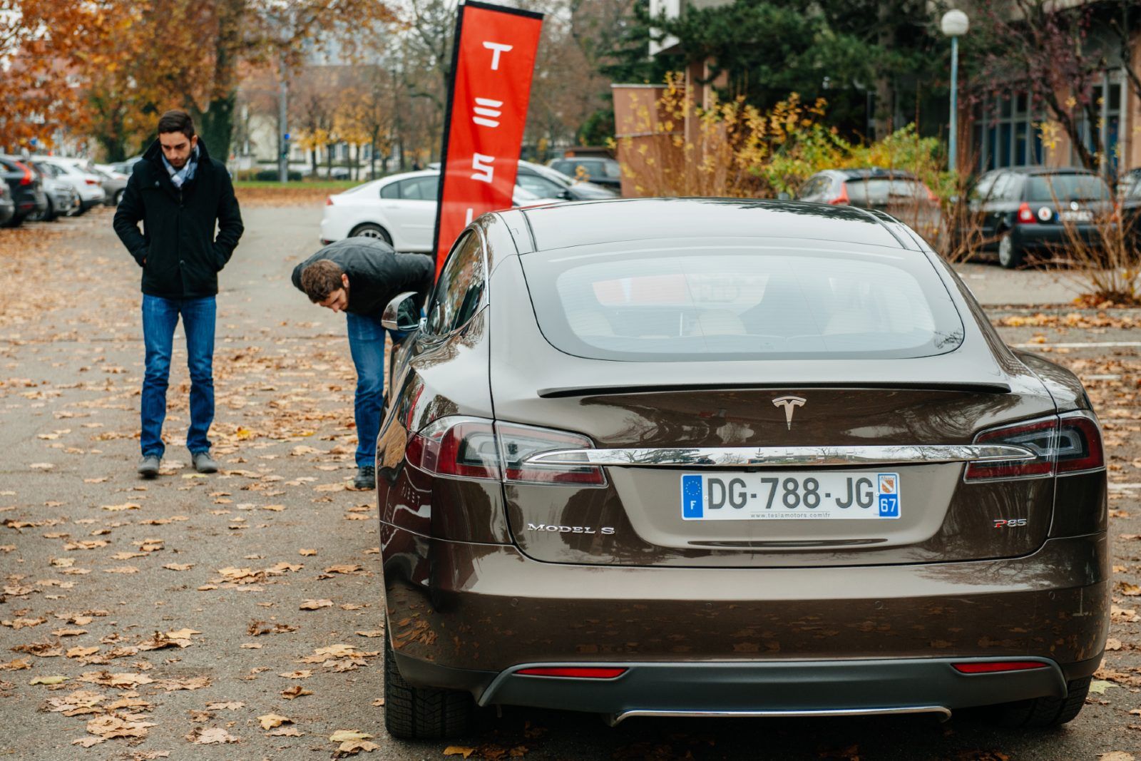 A brown Tesla Model S sits on a street in the autumn near a red Tesla flag - vehicle recalls