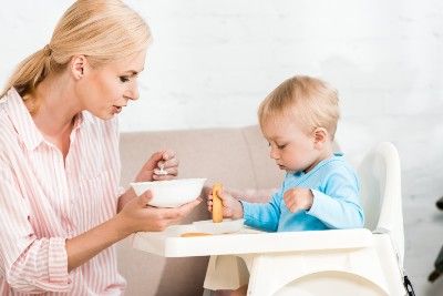 A blonde woman feeds a small child in a high chair - cuggl high chair recall