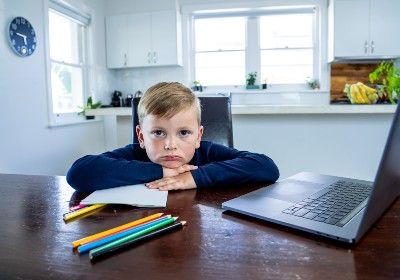 Young boy sitting with laptop and colored pencils looks sad at the camera - circuit breaker lockdown