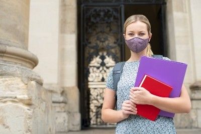 A female university student stands in front of a building while wearing a mask and carrying books - rights at university