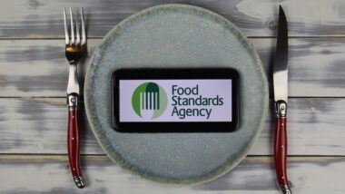 Closeup of smartphone with logo lettering of british food standards agency on plate with cutlery (focus on center of phone screen).