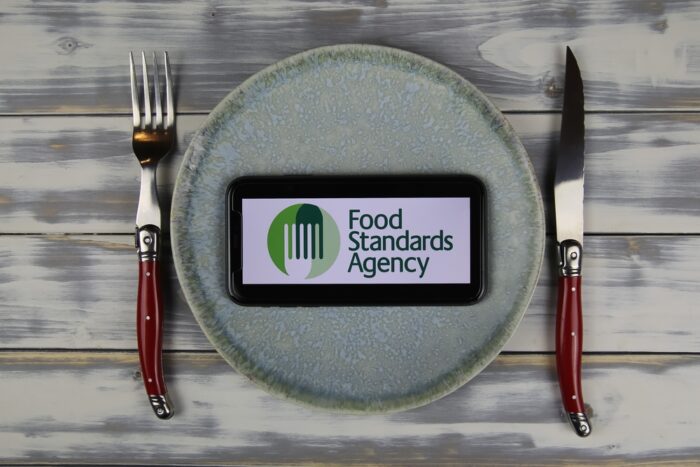 Closeup of smartphone with logo lettering of british food standards agency on plate with cutlery (focus on center of phone screen).