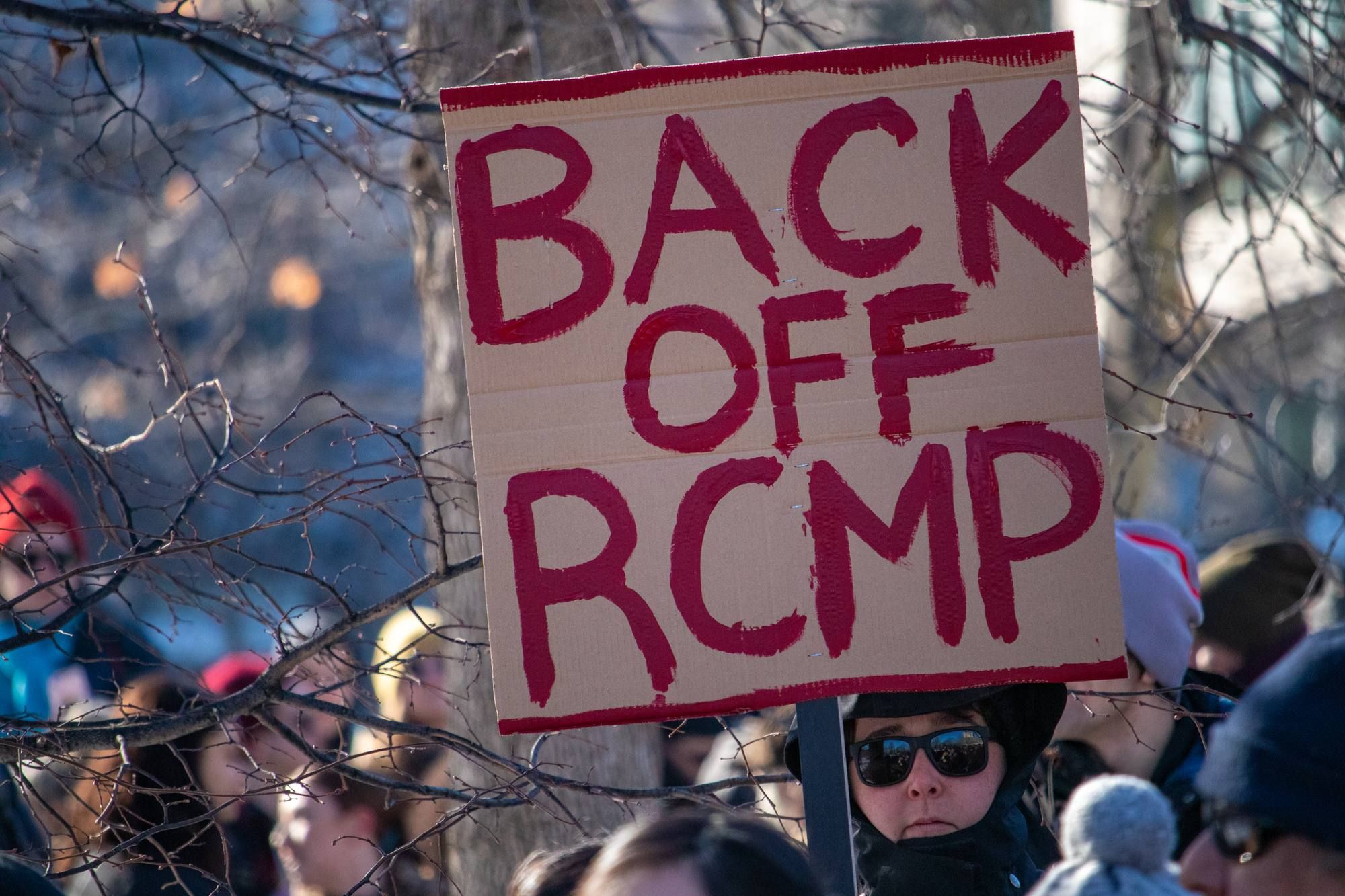 Person holding sign that reads Back off RCMP regarding the RCMP discrimination class action lawsuit expanding 