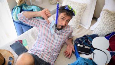 Guy gives a thumbs down while sitting back on his bed while wearing a snorkel, surrounded by open suitcase full of vacation items - td bank