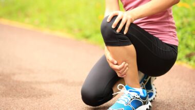 A female runner on a trail crouches down, grabbing her knee in pain
