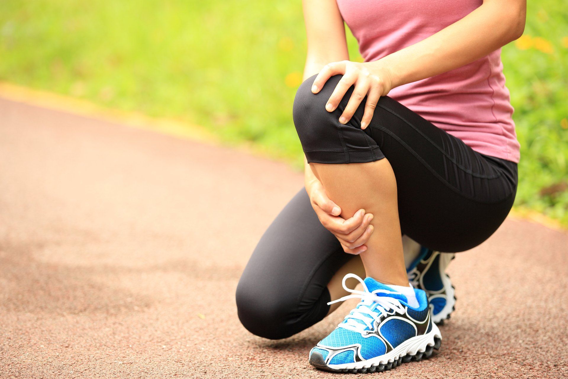 A female runner on a trail crouches down, grabbing her knee in pain
