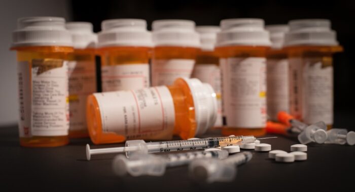 An injectable drug is loaded into a syringe while prescription medication is strewn about haphazardly, opioid epidemic concept.
