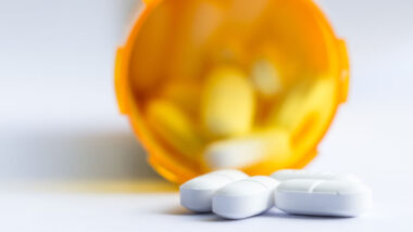 Close up of a group of white tablets with an out of focus prescription bottle in the background.