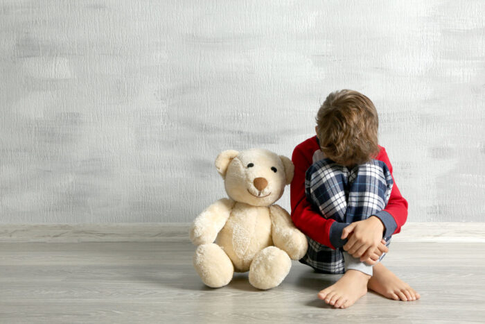 Sad little boy with teddy bear sitting on floor in empty room.